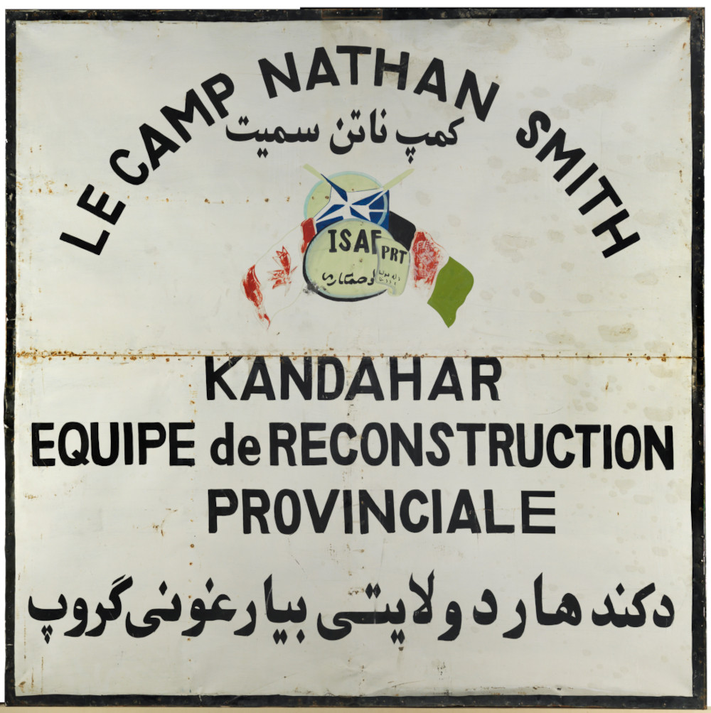 A square white sign with a very worn design of the Canadian and Afghanistan flags and large French and Arabic text reading “LE CAMP NATHAN SMITH KANDAHAR EQUIPE de RECONSTRUCTION PROVINCIALE.”