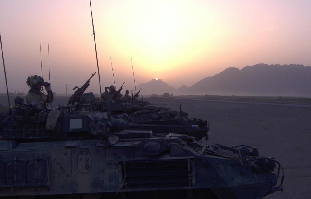 A line of armoured vehicles backlit by a hazy sun just above distant mountains.