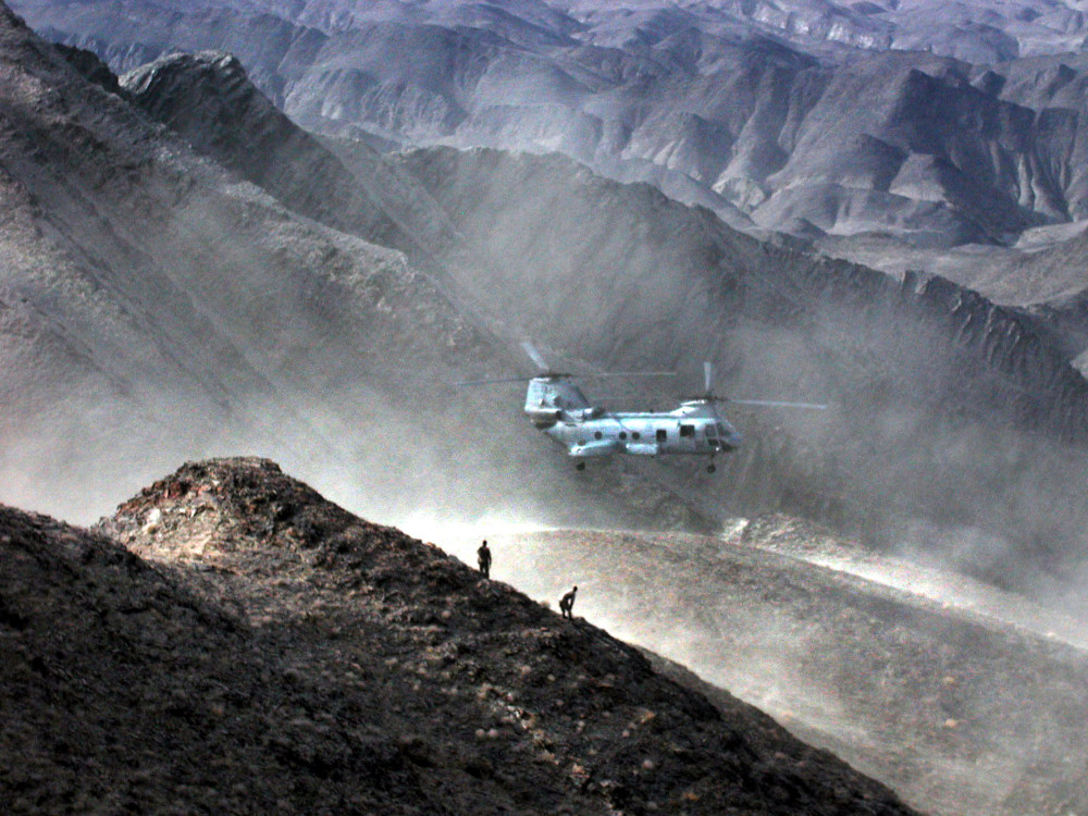 A large twin-rotor helicopter hovers amidst a mountainous landscape with two tiny human figures visible in silhouette on a ridgeline.
