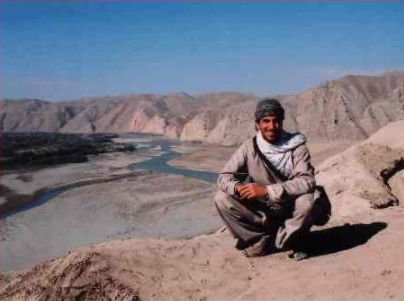 A smiling person with medium skin, a wool hat, and loose tan clothes crouches on a low mountain ridge, facing the camera, with a river valley in the background.]