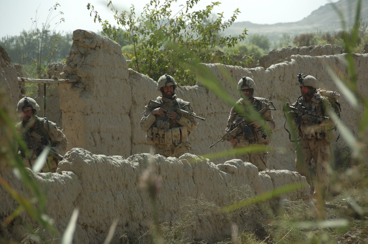 Four soldiers in tan camo carry weapons and walk between mud walls with greenery all around.