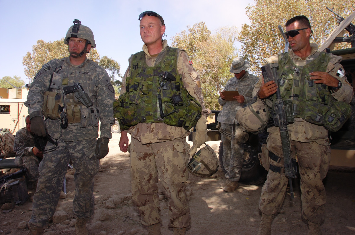 A soldier with short grey hair, tan camo, and a green vest stands between a soldier in grey camo and a helmet (on the left) and a soldier with very short dark hair, sunglasses, tan camo, a green vest, and a firearm (on the right).