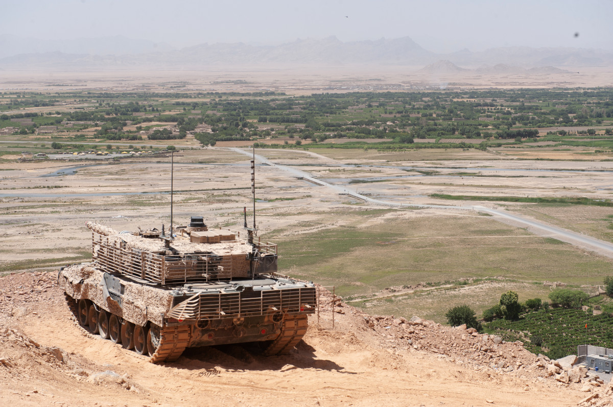 A tan camo military tank sits on a height above a very wide, flat green landscape, with a ridge of mountains in the far distance.