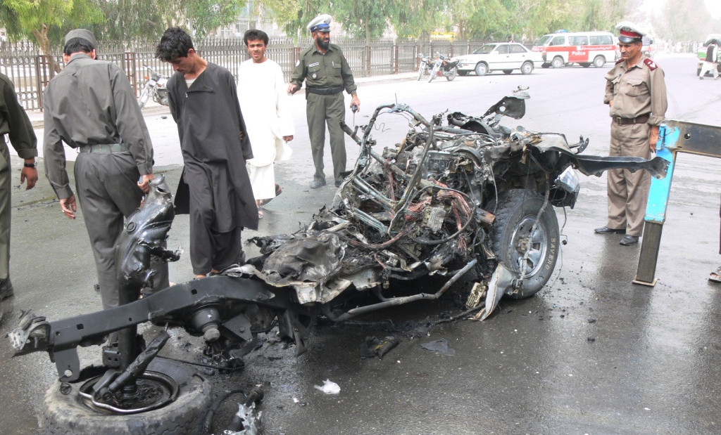 Several onlookers stand in a street examining the burned and twisted chassis and wheels of what might have been a car.