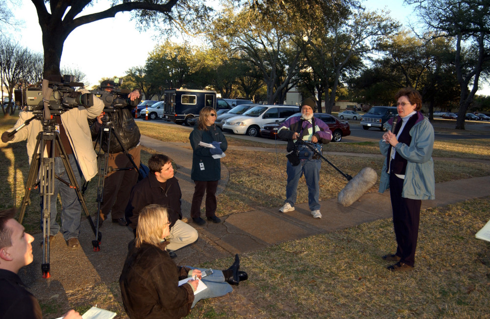 A person stands and speaks in front of a crowd of reporters.