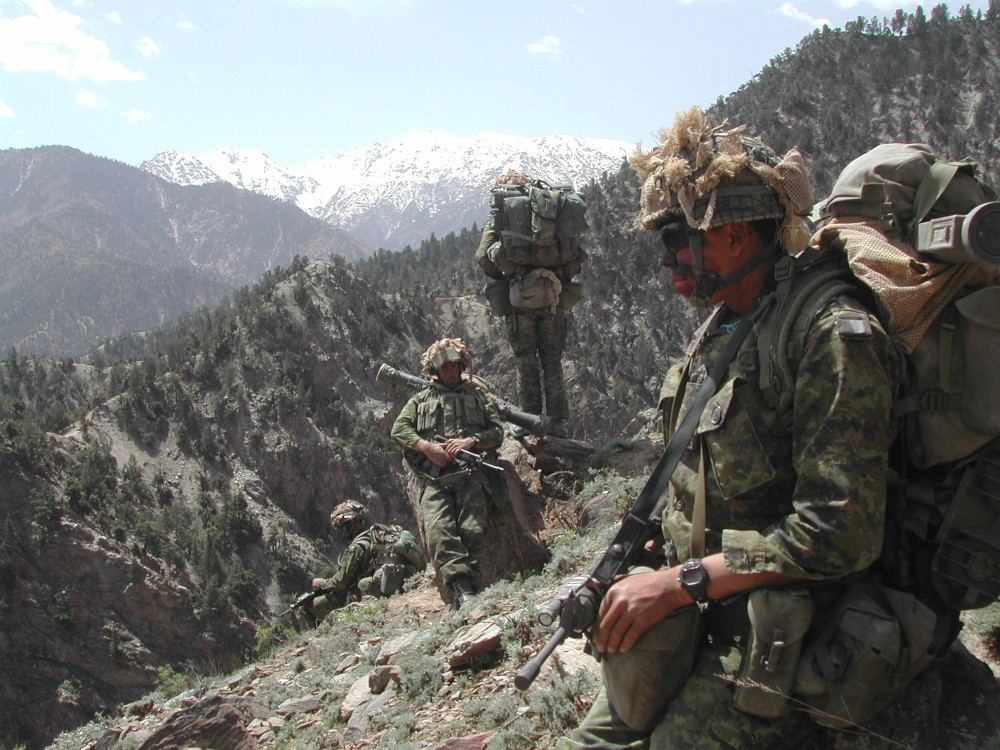 Four soldiers in green camo and face paint gathered on a slope with near and far mountain ridges behind them
