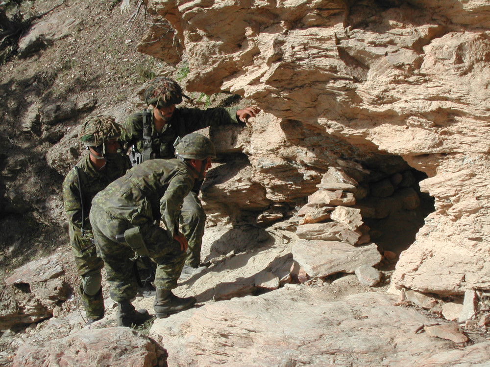 Three soldiers in green camo peer into a small cave entrance.