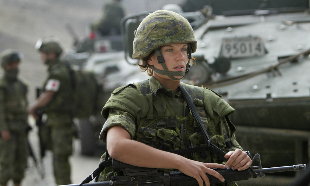 A female soldier in green camo and a helmet, holding a weapon, walks next to some large, armoured vehicles, with more soldiers visible behind her.
