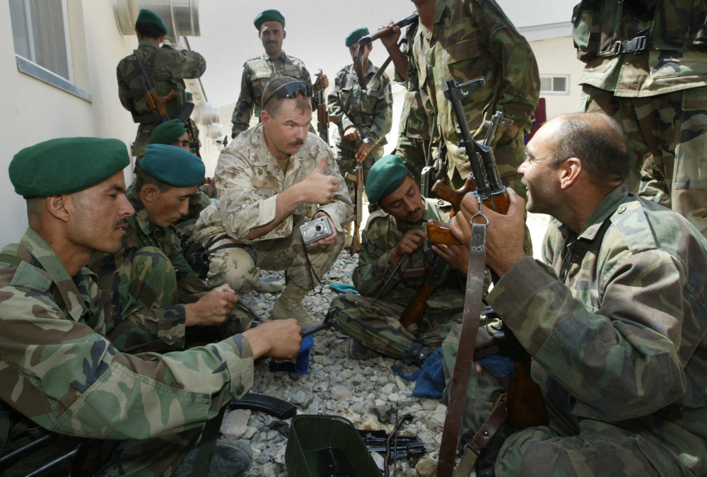 A light-skinned soldier with a moustache and tan camo uniform squats and gives a “thumbs up” gesture amongst a group of medium-dark-skinned soldiers in green camo who are holding or working on submachine guns.