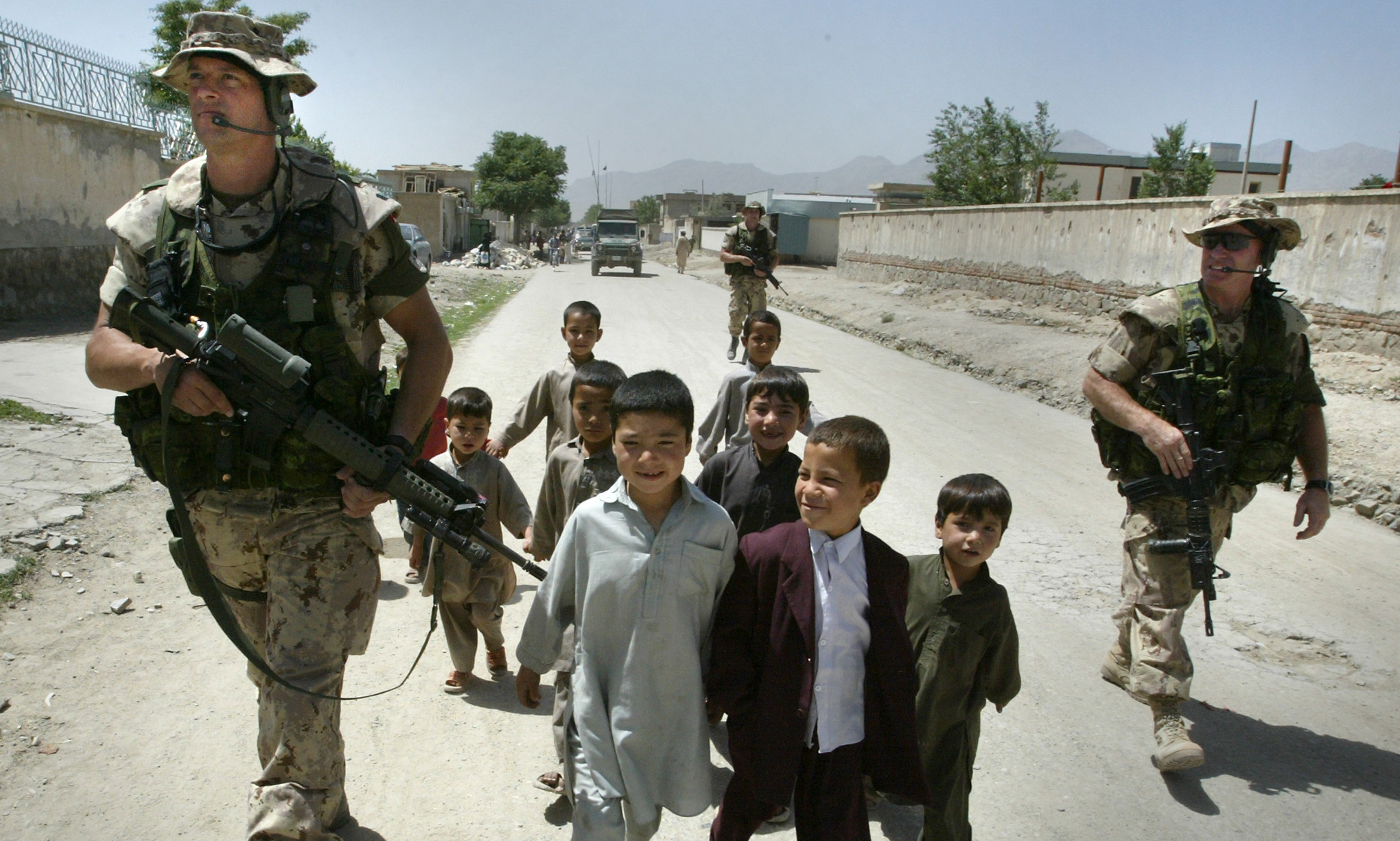 Three soldiers in tan camo and carrying weapons walk down a dirt road lined with mud walls as a crowd of small children walk with them.