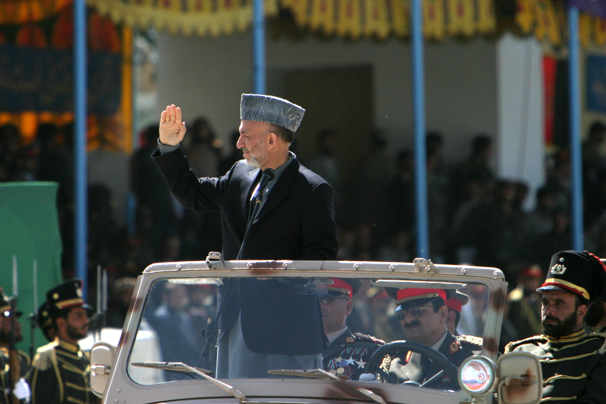 A medium-skinned man with a short greying beard, a grey hat, and a dark jacket stands in a large open-top car and waves, surrounded by seated men in highly decorated uniforms.