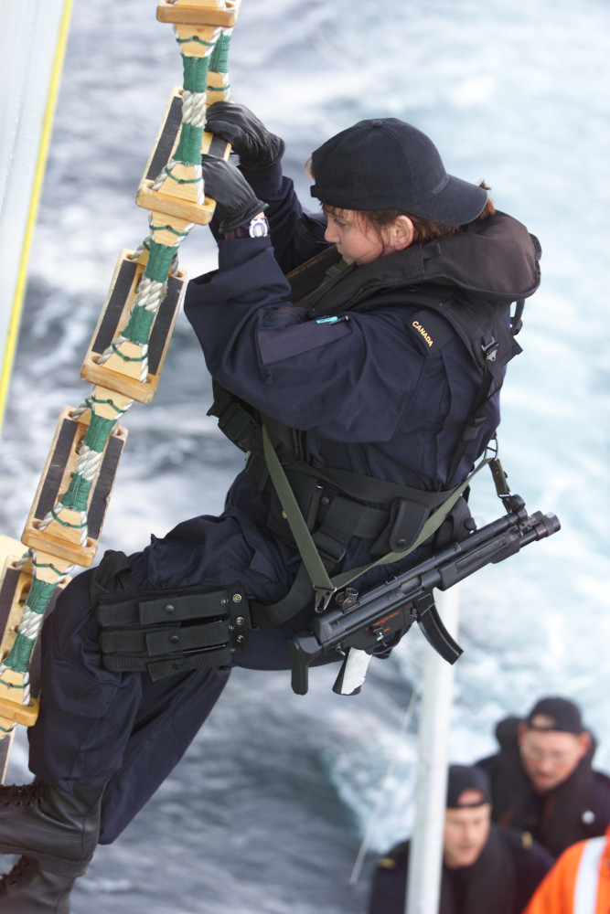 A person with a dark blue uniform and a submachine gun slung across their body climbs a steep rope ladder above a body of water, with people in a small boat partially visible below.