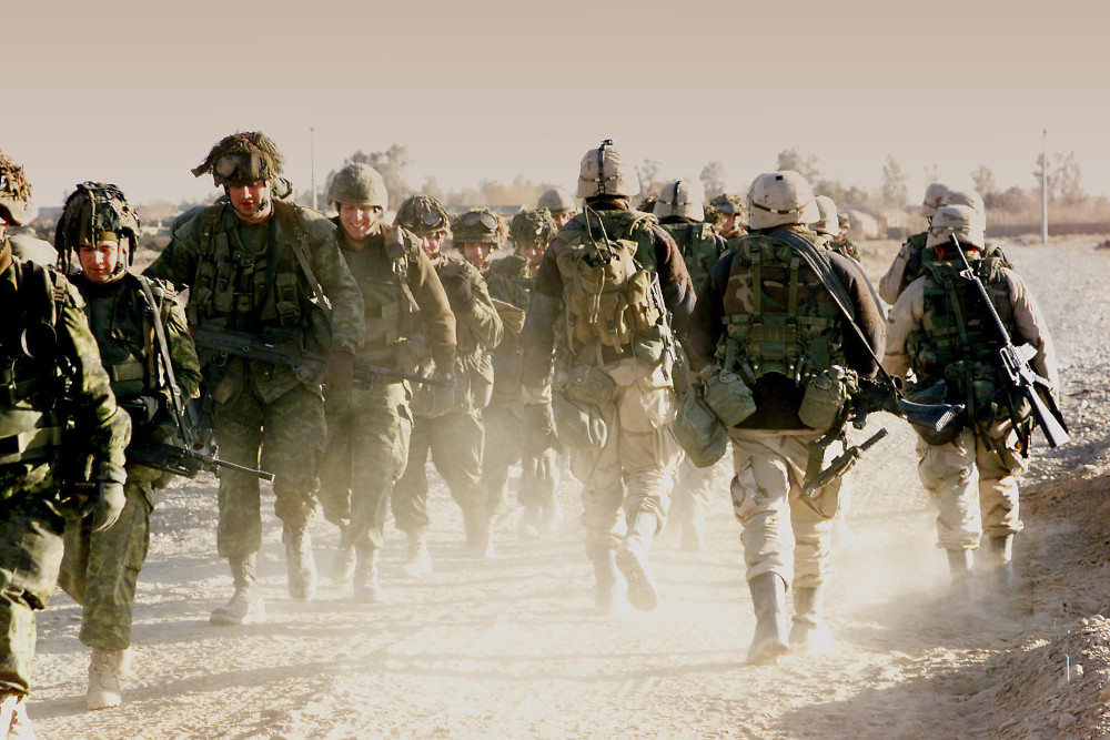 A group of soldiers wearing green camo uniforms walks towards the camera, passing another group in tan camo uniforms walking the other way.