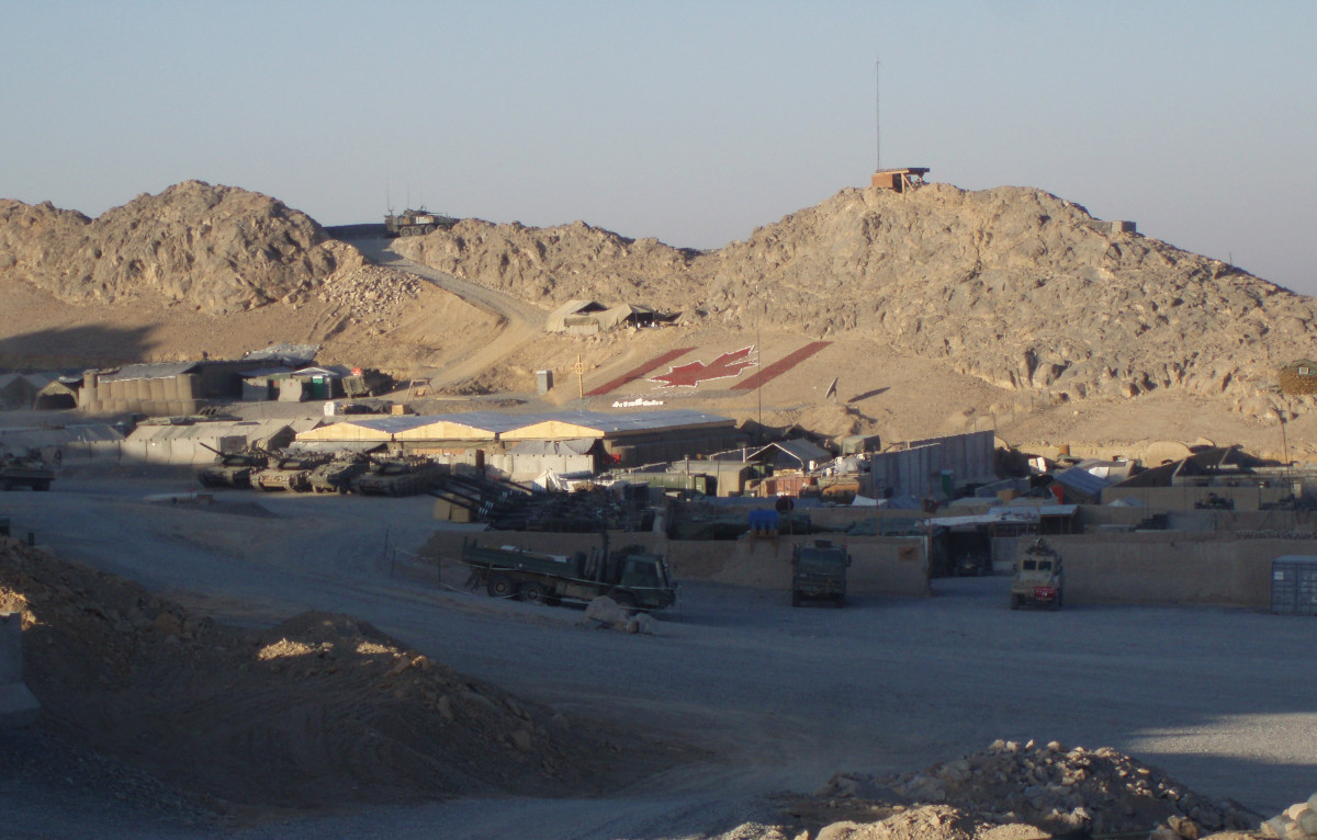 [Some low brown buildings and green military vehicles with rocky hills in the background. A very large Canadian flag design has been created on a hillside behind the buildings.