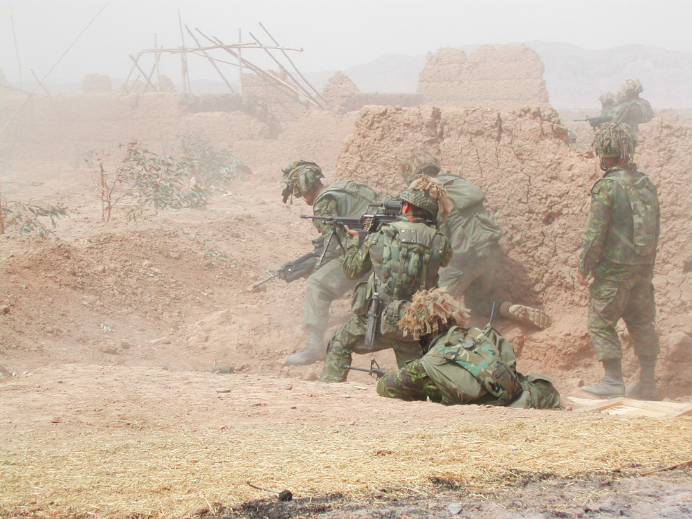 A group of several soldiers in green camo hold weapons as they shelter and peer around a mud wall, with more mud and wood structures visible in the distance.
