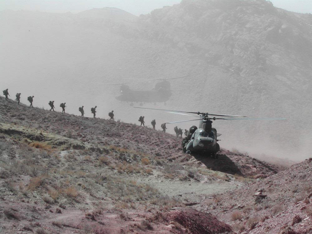 A line of soldiers seen in silhouette walking down a ridge line towards a large helicopter, with another helicopter hovering further away and a mountain in the distance.