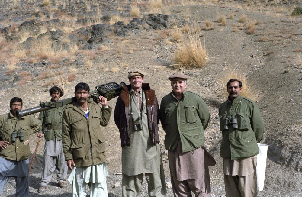 A light-skinned man in a brown bomber jacket stands with five medium-dark-skinned men wearing green jackets, one carrying a large weapon across his shoulders.]