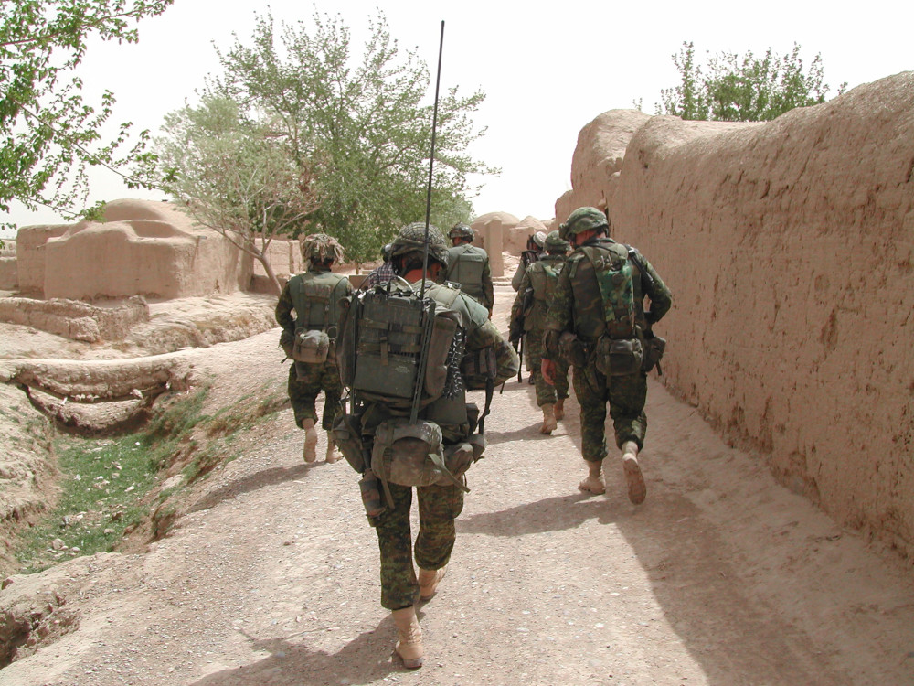 A group of soldiers in green camo walk away on a dirt road amongst tan coloured walls and structures.