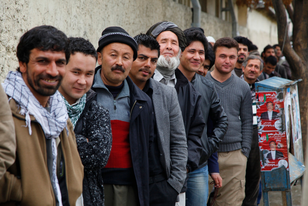 A long line of medium-skinned people in casual clothes stand along a wall, smiling at the camera.