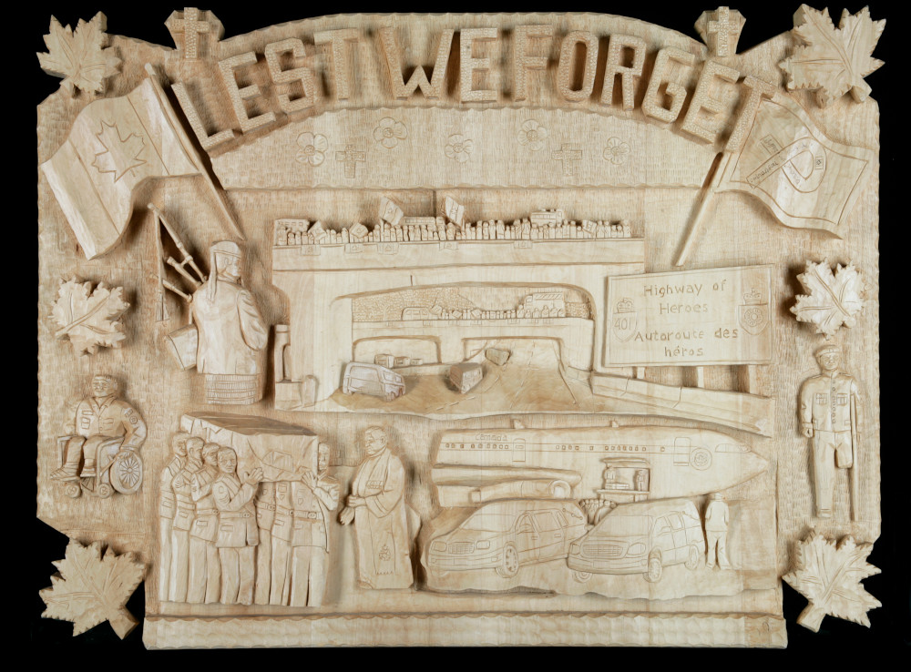 A wooden relief sculpture reading “Lest we forget” across the top and showing scenes from the repatriation of a soldier’s remains, including a group of soldiers carrying a casket and a crowd gathered on a highway overpass.