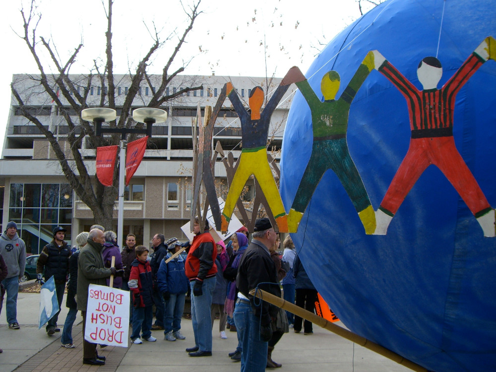 A group of people gathered in a public square, with a large sculpture of a globe surrounded by linked human figures in the foreground.