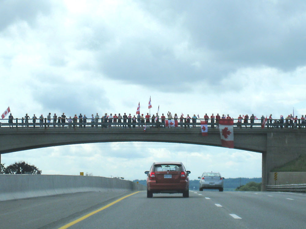 Cars drive on a highway beneath an overpass full of people, some holding large Canadian flags.