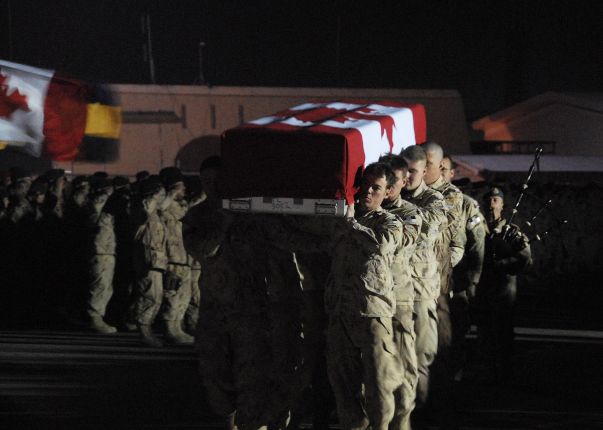 At night and brightly lit from the right, eight soldiers in tan uniforms walk toward the camera carrying a rectangular box draped with a large Canadian flag on their shoulders, with a bagpiper, more soldiers, and another flag behind them.