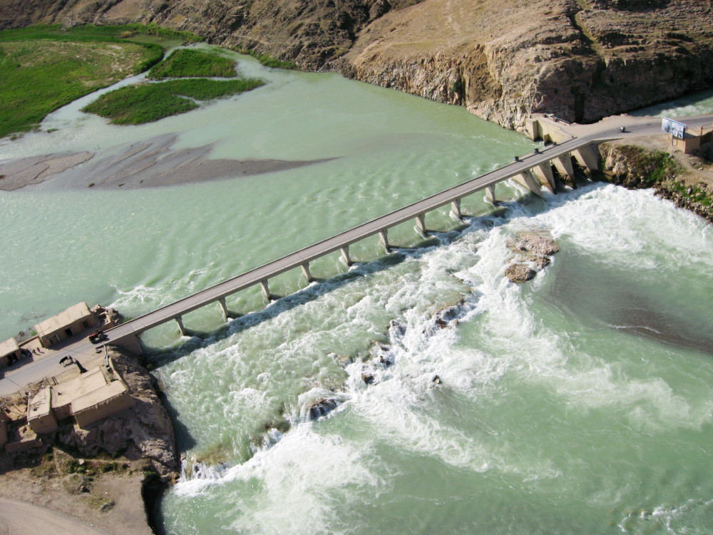 A narrow bridge spans a wide body of blue-green water flowing over low rapids.