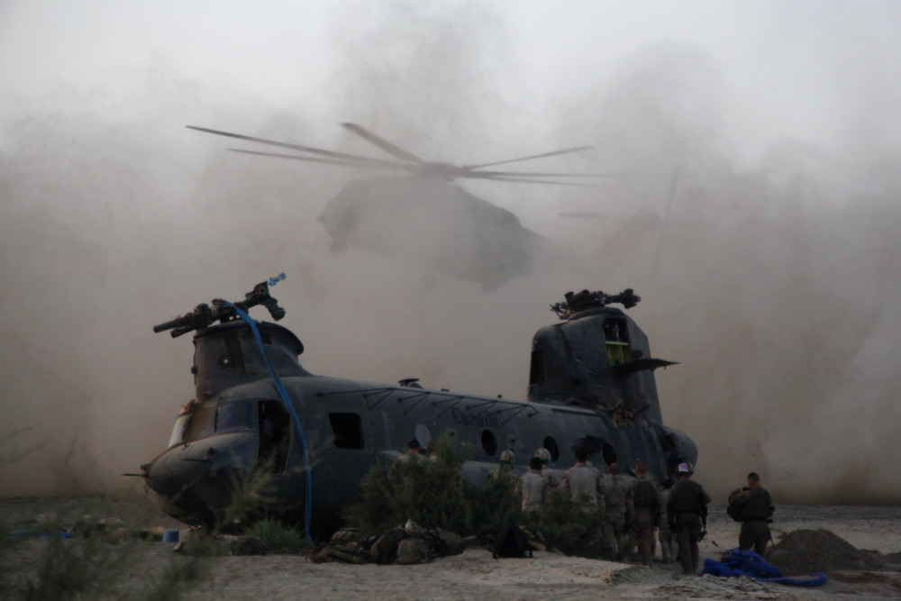 A group of soldiers stand in front of a large helicopter as another helicopter lands behind it in a huge cloud of dust.