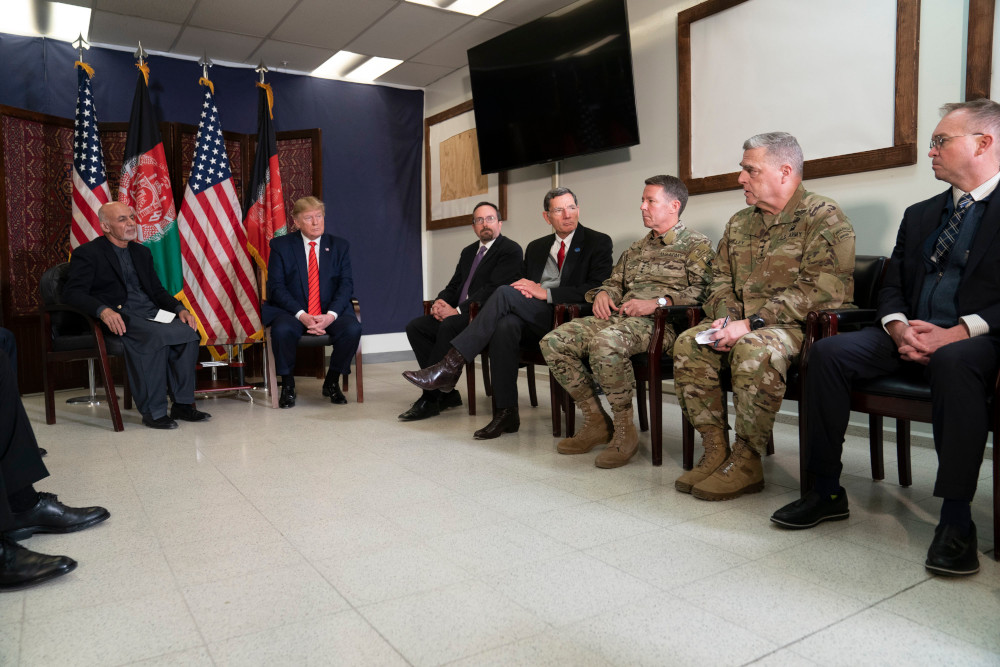 Several men, some in military uniforms and others in suits, sit in chairs around the perimeter of a room.