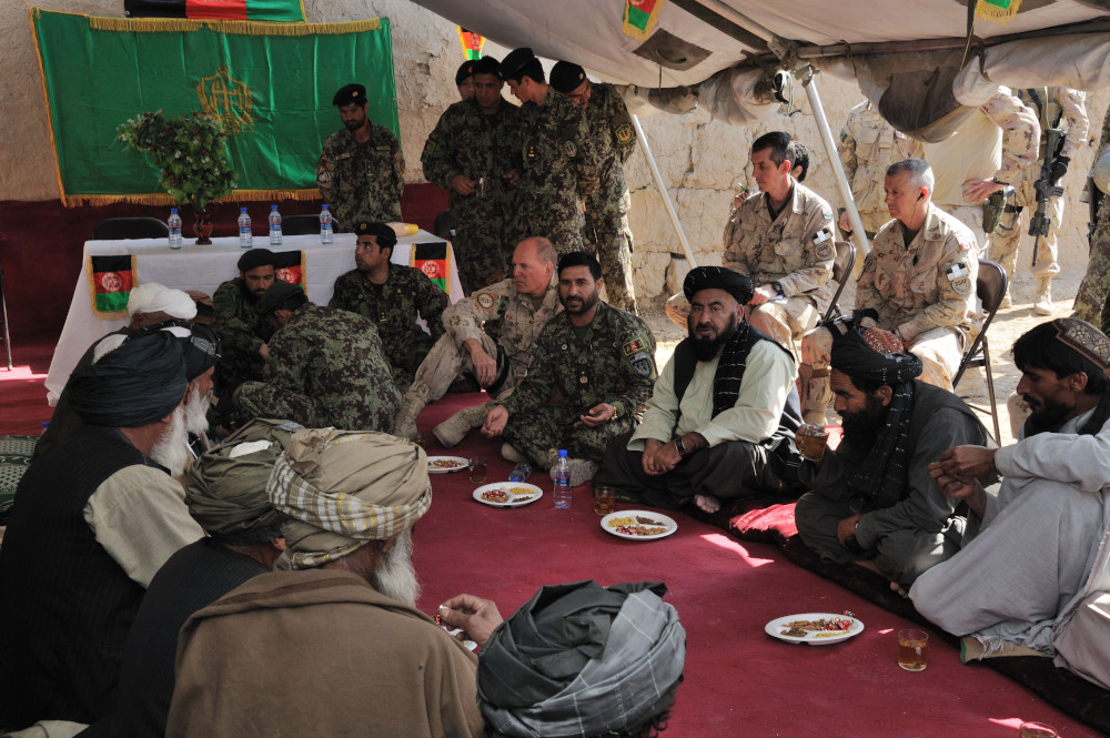 A group of men in both western military clothing and traditional Afghan robes sit on a red carpet, many with plates of food and glasses before them.