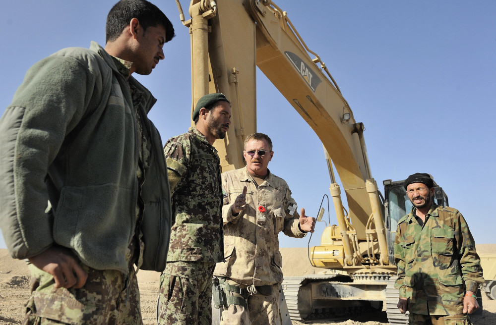 A light-skinned man in a tan combat uniform wearing a red poppy speaks with three medium-dark-skinned men in green uniforms, with a large construction vehicle in the background.