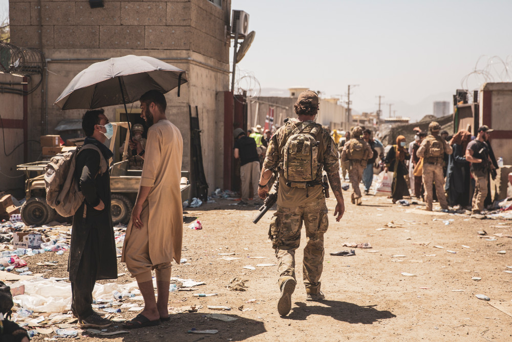 A soldier in camo walks towards a large crowd of soldiers and civilians amongst walls topped with barbed wire.