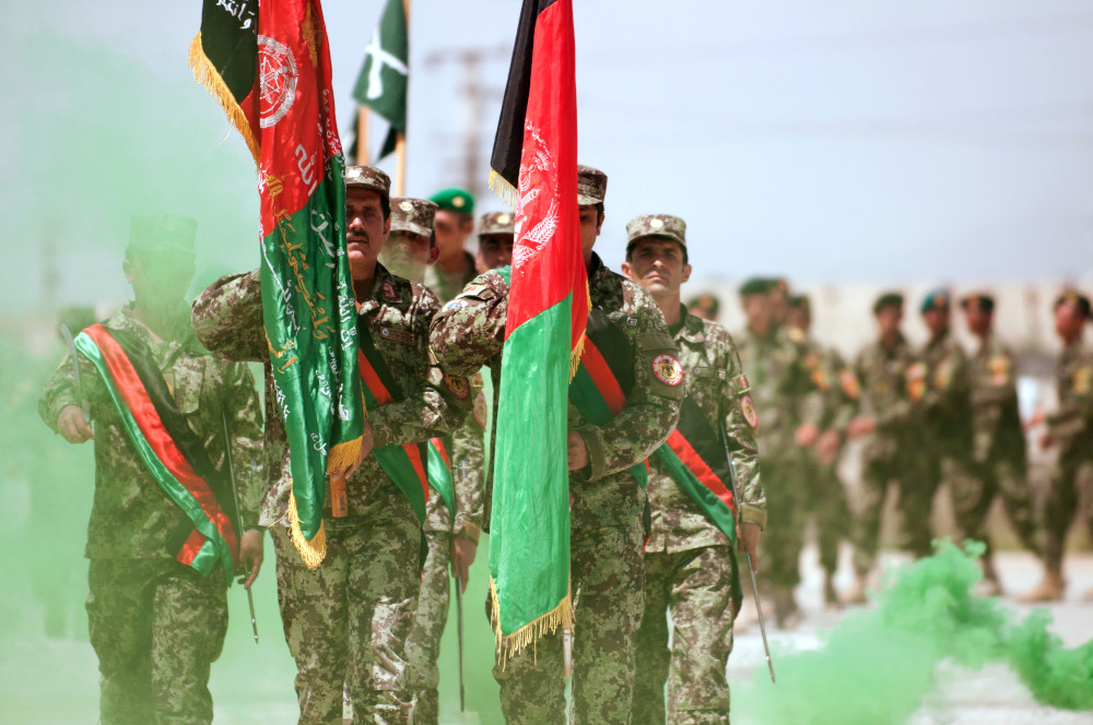A group of medium-dark-skinned people in camo uniforms march carrying Afghanistan flags.