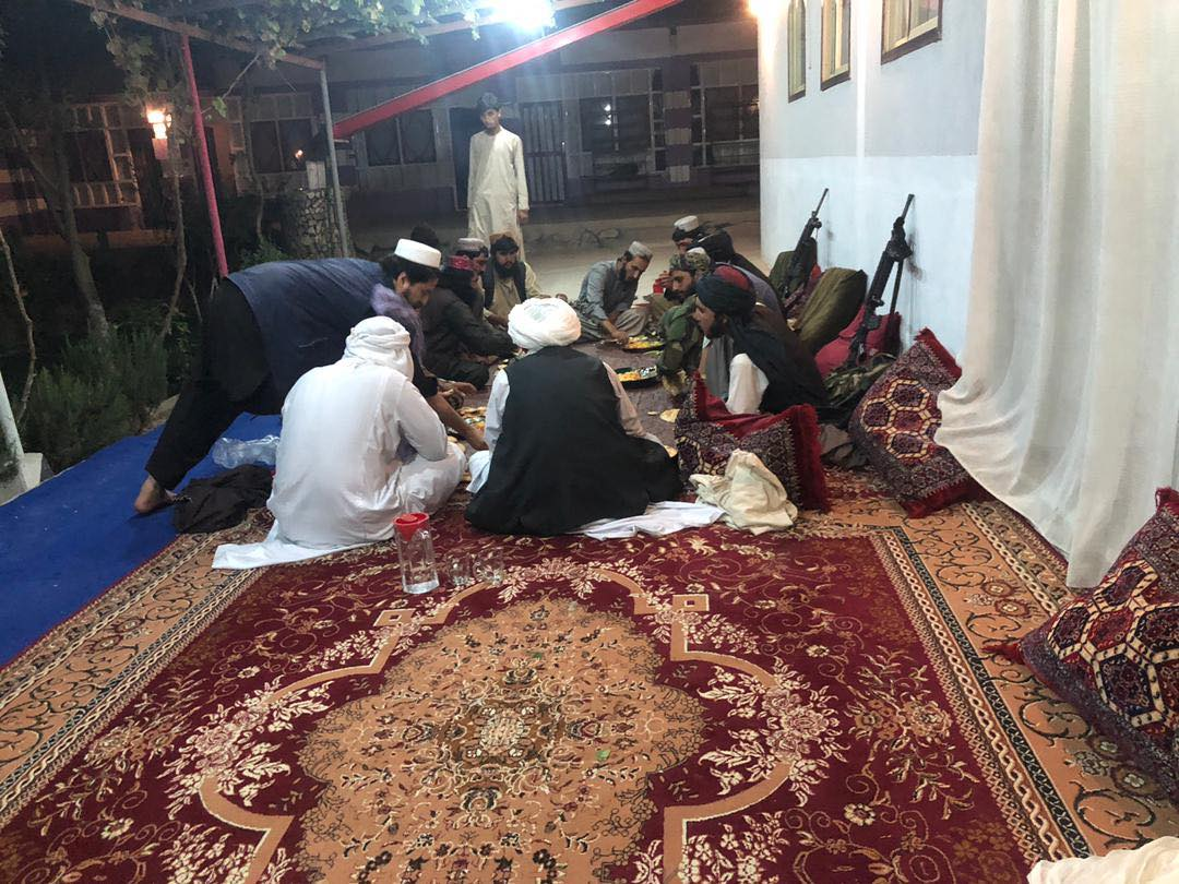 A group of medium-skinned people in flowing clothes and head coverings sit on a carpeted patio near a row of pillows and automatic weapons leaning against a wall.