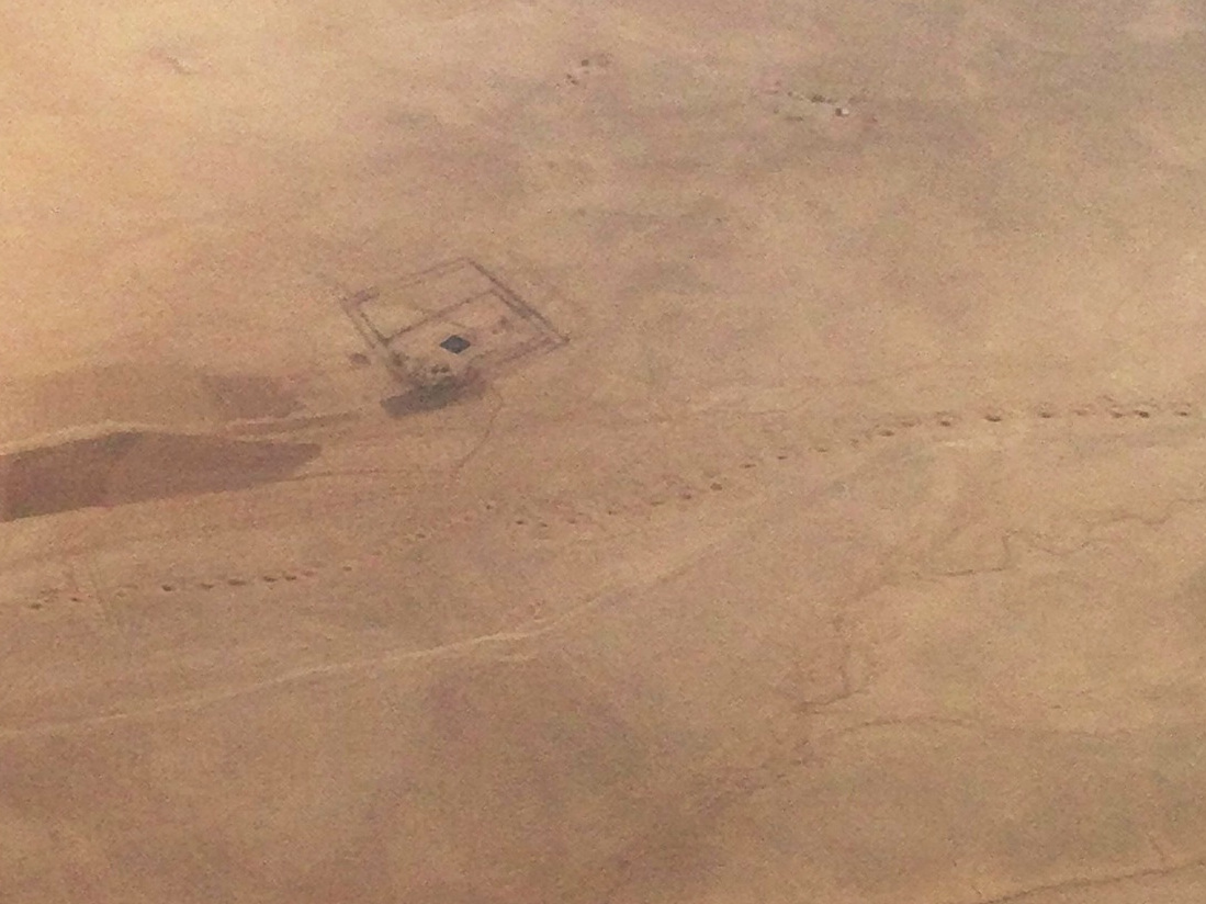 Aerial photo of an arid landscape showing a road and other regular structures, wandering riverbeds, and a linear series of dots extending the width of the photo.