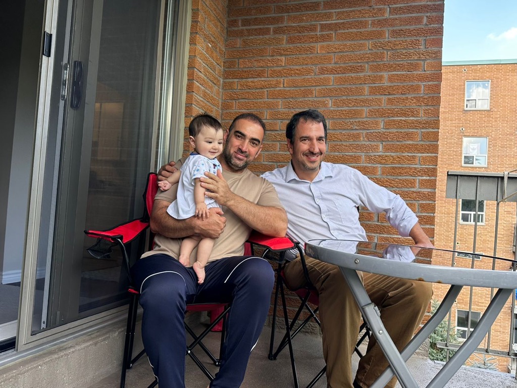 A smiling medium-skinned man holds an infant and sits next to a smiling light-skinned [AB1.1]man, on an apartment’s balcony.