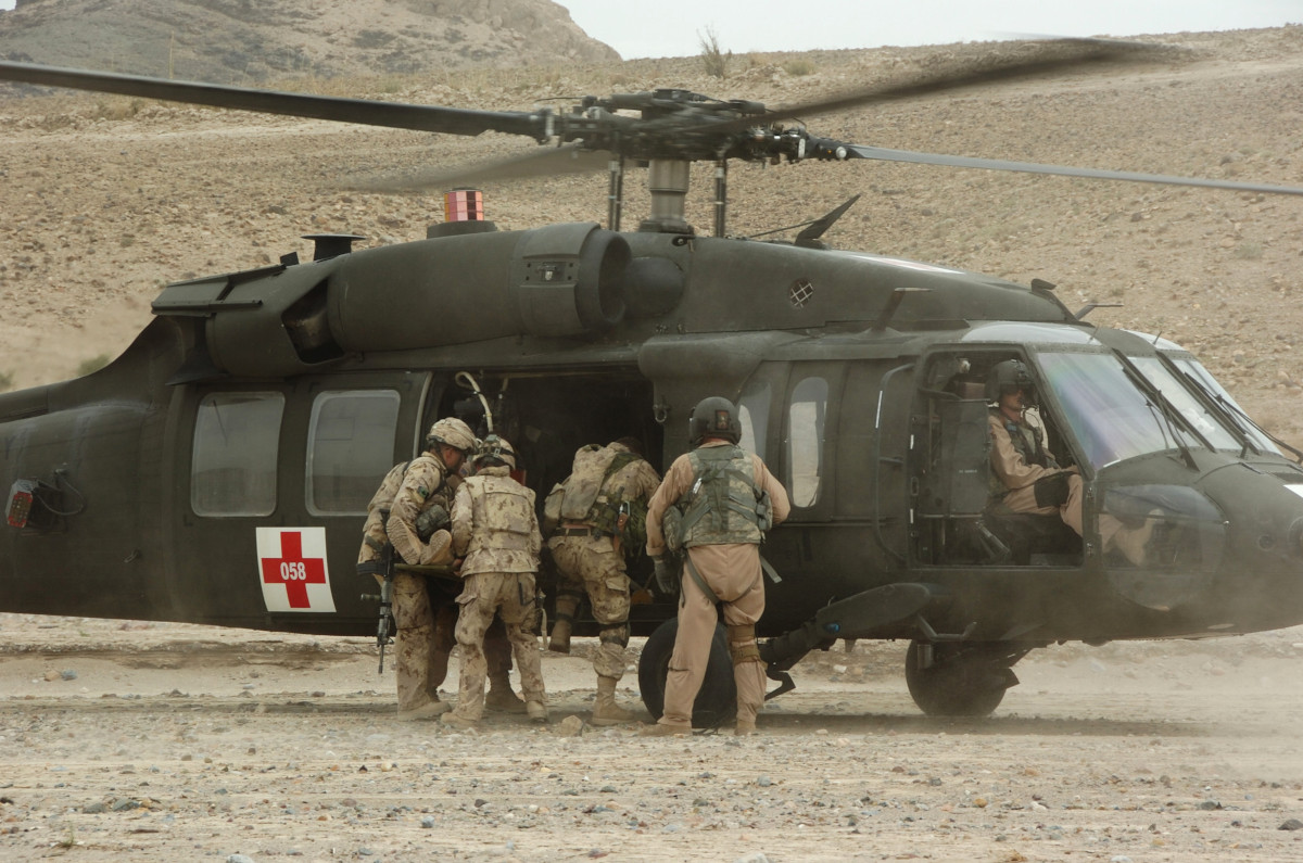 A group of soldiers in tan camo climbing aboard a dark green helicopter marked with a red cross.