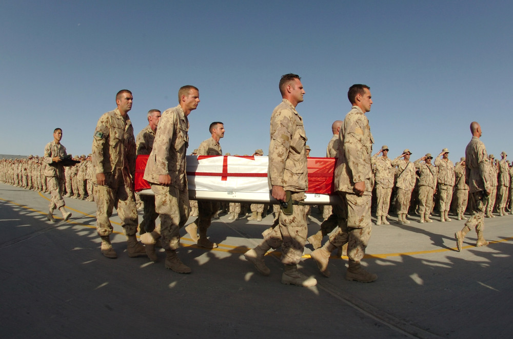 A procession of soldiers in tan camo uniforms carry a metal coffin draped with the Canadian flag past a long line of soldiers saluting.