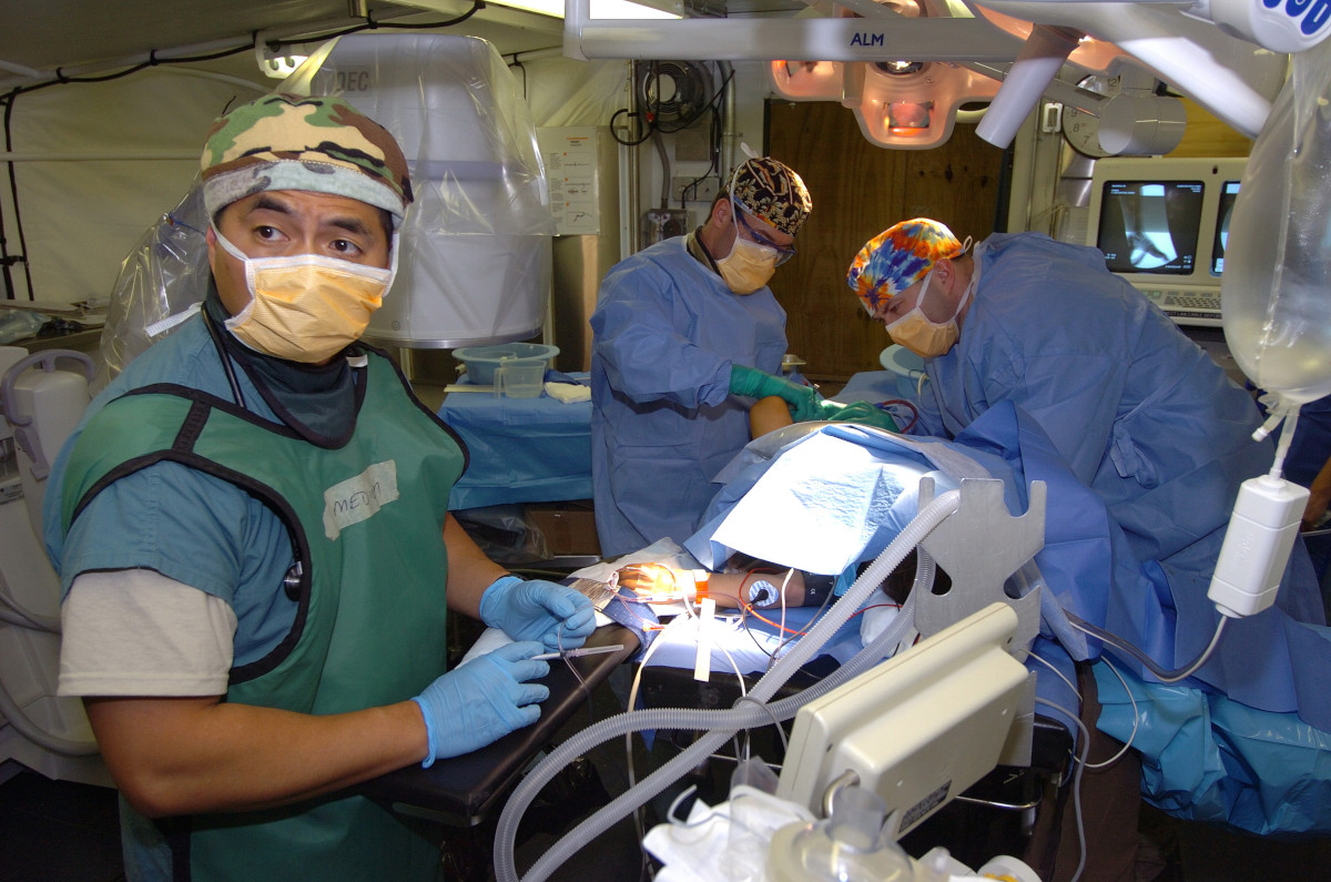 In the foreground, a medium-skinned person wearing medical scrubs and a mask looks back towards the camera, as two more people in scrubs and masks operate on someone.