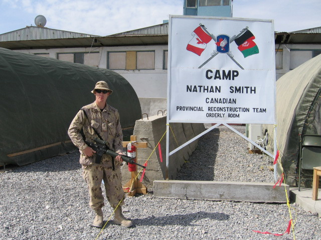 A soldier with a tan uniform and firearm stands next to a large white sign with crossed Canadian and Afghanistan flags, reading “Camp Nathan Smith Canadian Provincial Reconstruction Team.”