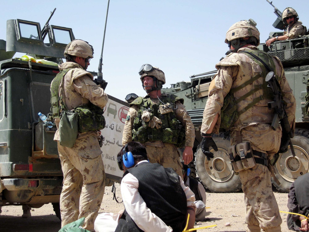 Three armed soldiers in tan camo uniforms stand near two large green armoured vehicles and three people in civilian clothes who sit on the ground wearing blindfolds and blue plastic earmuffs with their hands bound behind them.