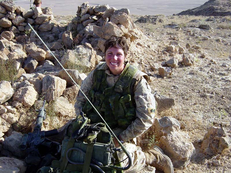 A smiling soldier with short wavy hair, tan camo uniform, and a green ballistic vest sits in a rocky landscape.
