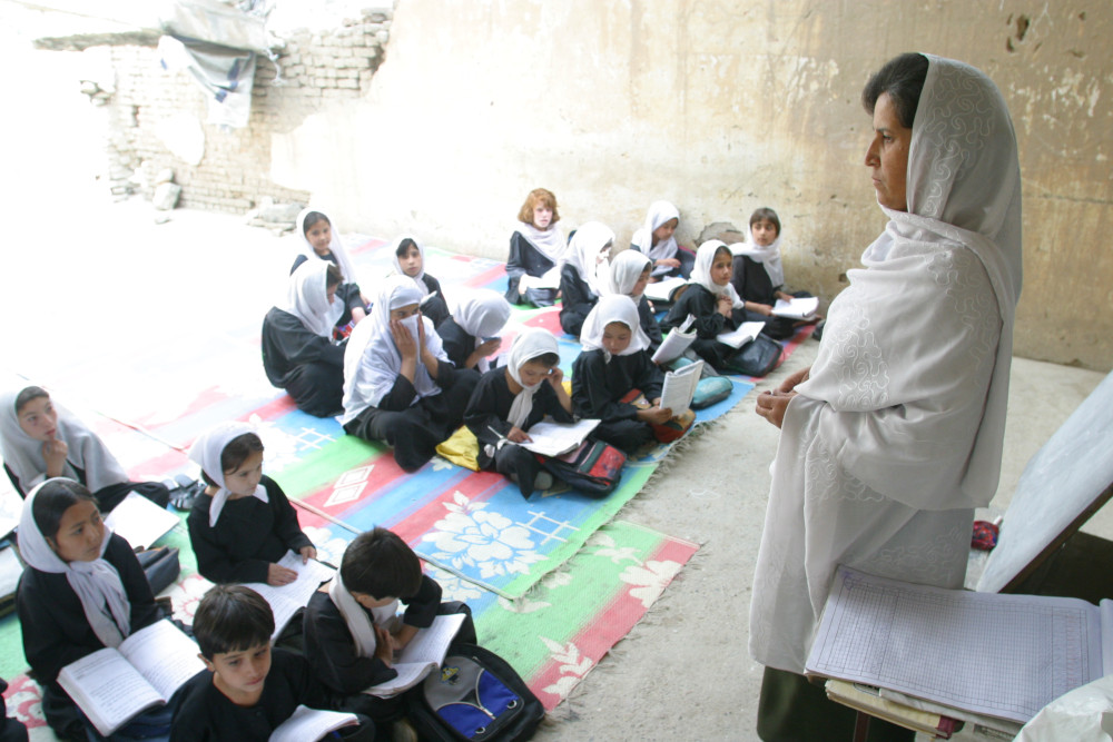 A teacher wearing a long headscarf speaks to a class of children on the floor, most of them also in headscarves.
