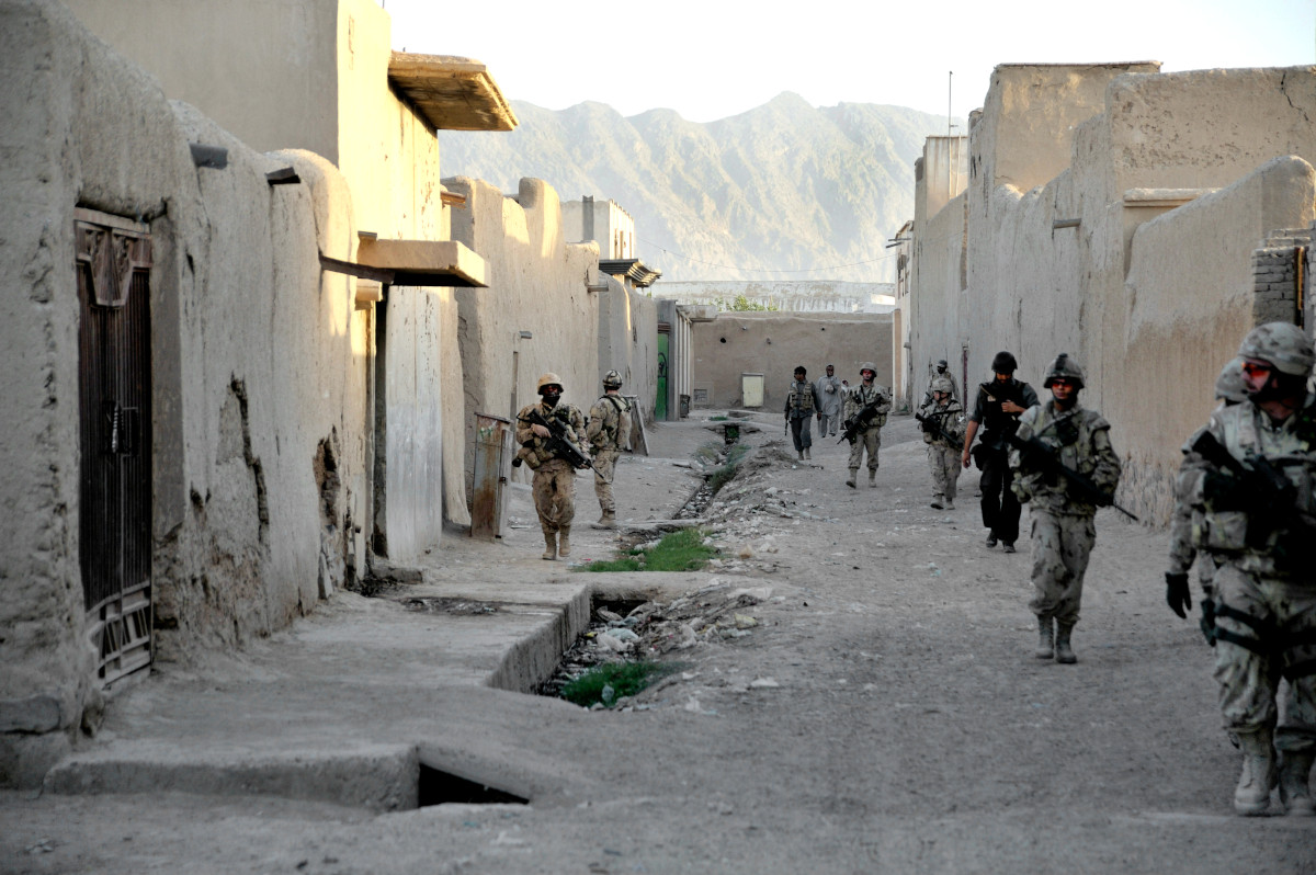 Several armed soldiers walk down both sides of a street lined with low buildings and walls.