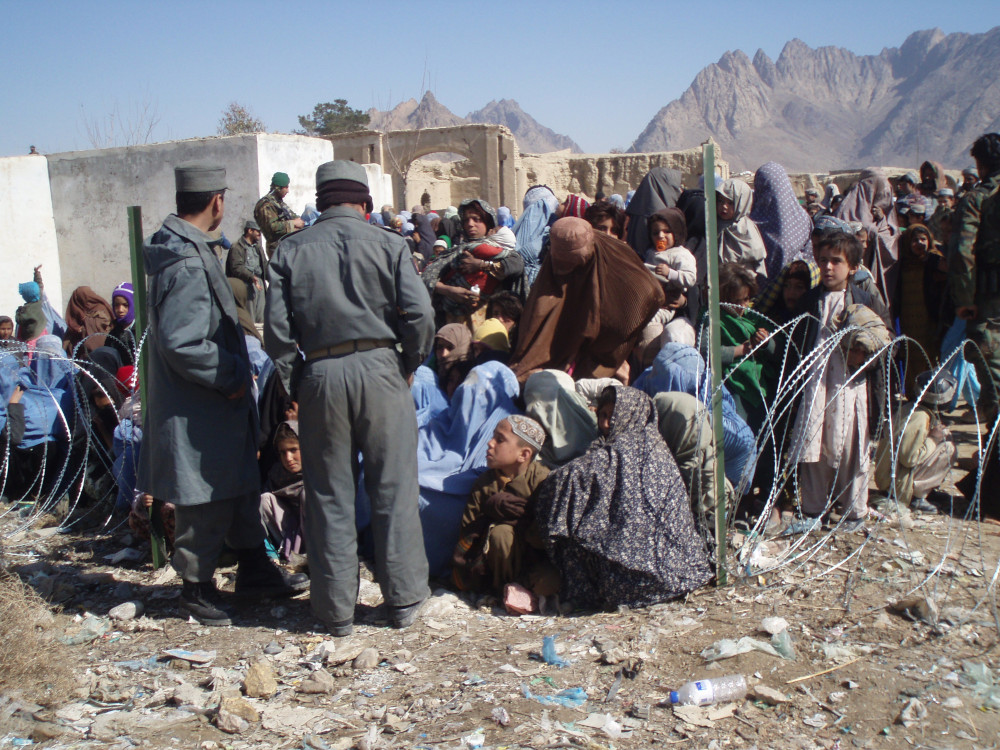 A large crowd of people, many wearing flowing clothes and head coverings, stand and sit near some low mud buildings, as two people in blue-grey uniforms stand nearby.