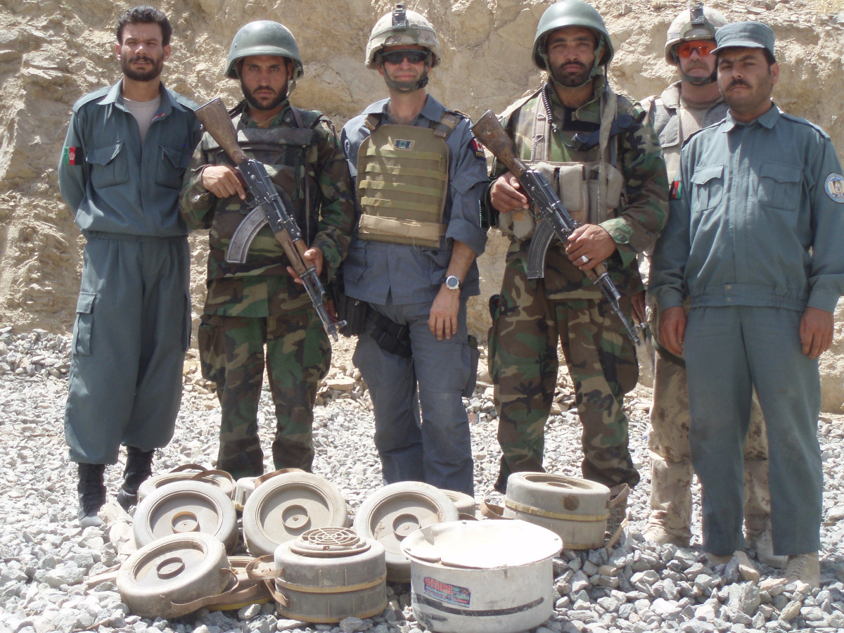 Seven people, some in blue uniforms and some in green or tan camo, stand in front of a dozen or so short cylindrical objects including landmines and pots.