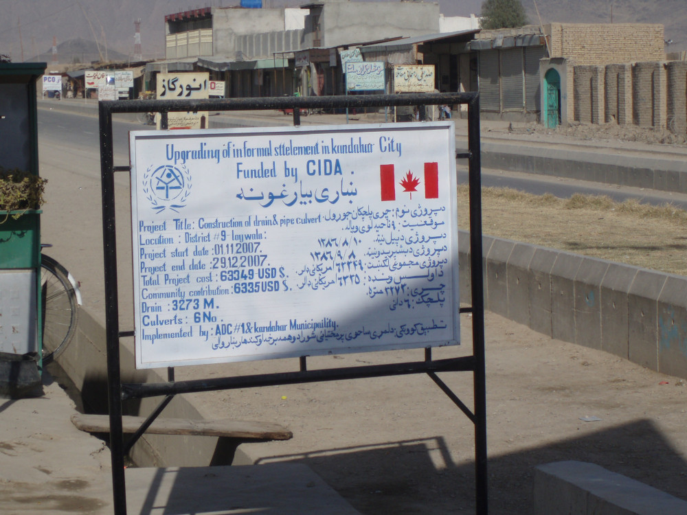 A roadside sign in English and Pashto with the large title “Upgrading of informal settlement in Kandahar City funded by CIDA” above lists of information in much smaller text.