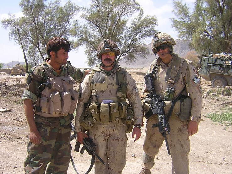 A medium-dark-skinned soldier in green camo stands next to two light-skinned soldiers in tan camo with trees and armoured vehicles visible in the background.