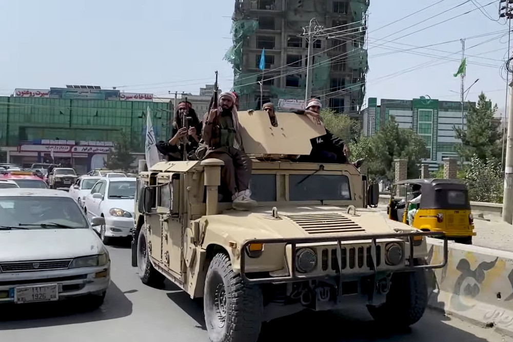 Armed men sit atop an armoured vehicle driving in urban traffic.